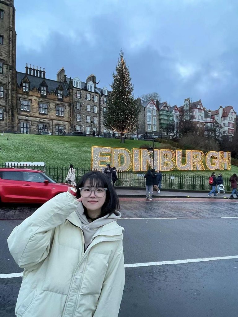 Rongjie Wang poses in front of sign reading, "Edinburgh" in England