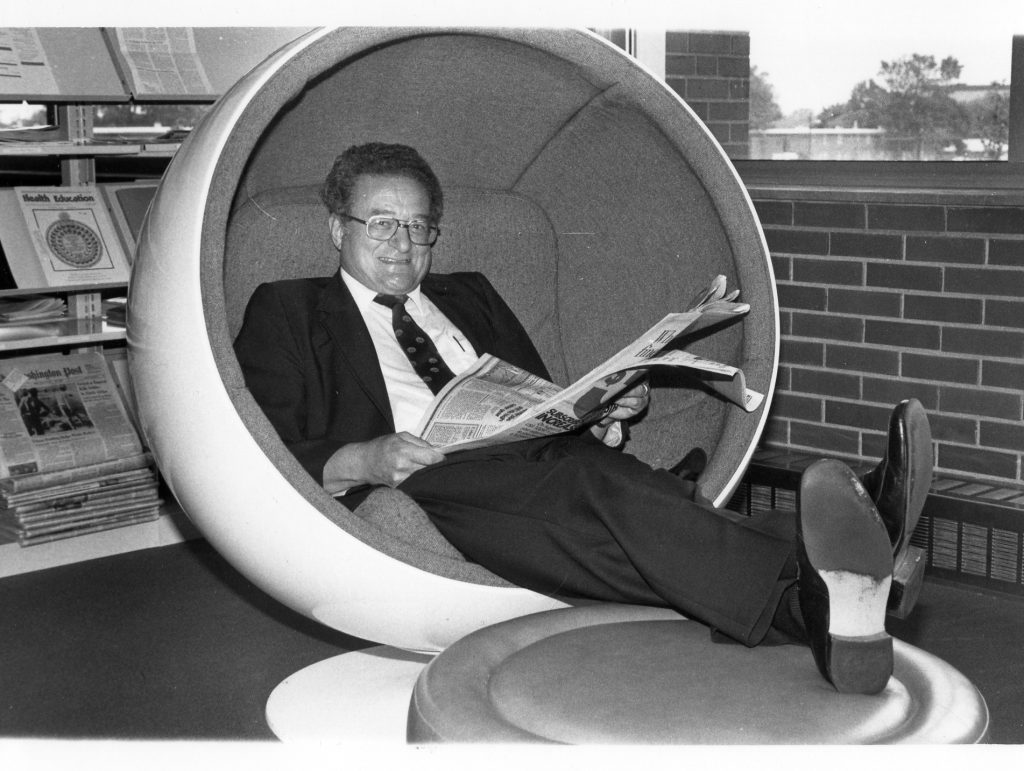 Black and white photo of Dr. Gene Sease reading a newspaper in a UIndy egg chair