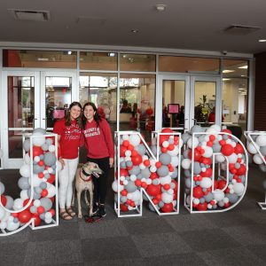 Photo of Grady the Greyhound with two UIndy students standing with the balloon sign that spells "UINDY"