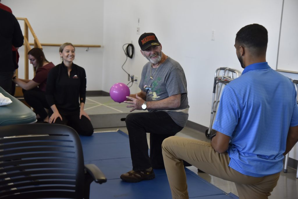 A community patient resource group volunteer works with a purple medicine ball under supervision of UIndy physical therapy students