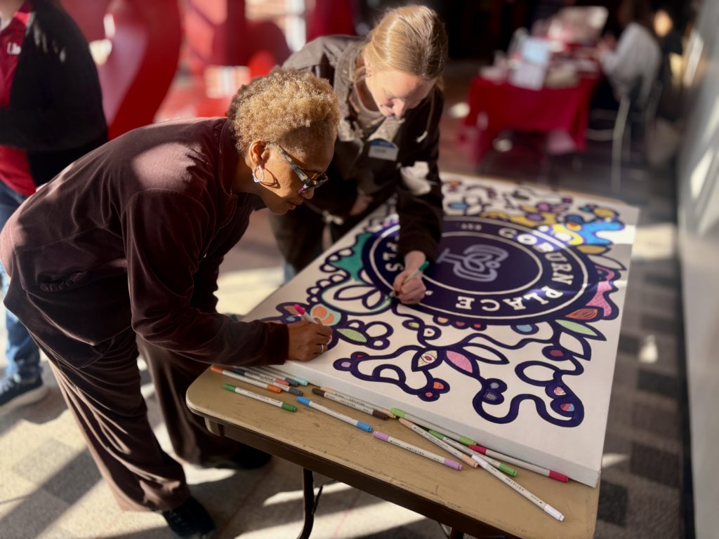 Two women lean over a table coloring a large decorative sign with markers.