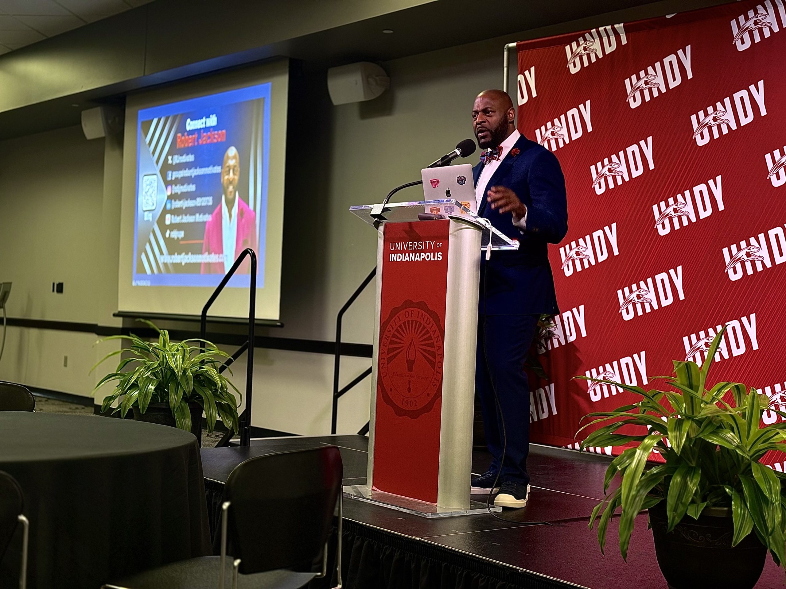 A man in a navy suit speaks at a podium on a stage at the University of Indianapolis, with a red UIndy backdrop behind him