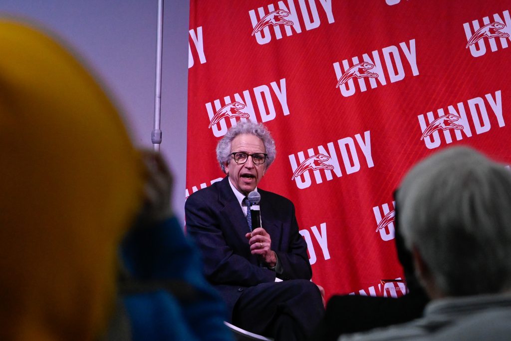Man on stage holding a microphone with UIndy backdrop, speaks to packed room.
