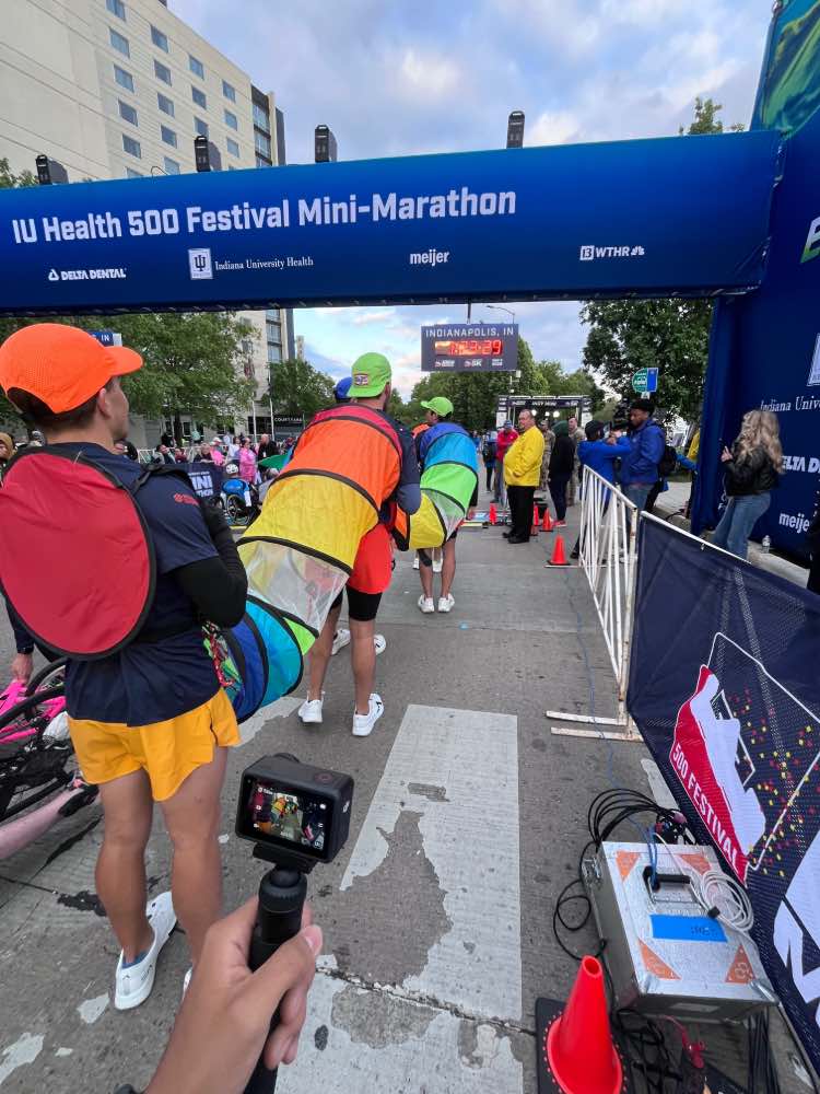 Five men in caterpillar costume line up at starting point of race with IU Health 500 Festival Mini Marathon banner above. Spectators watching in background.