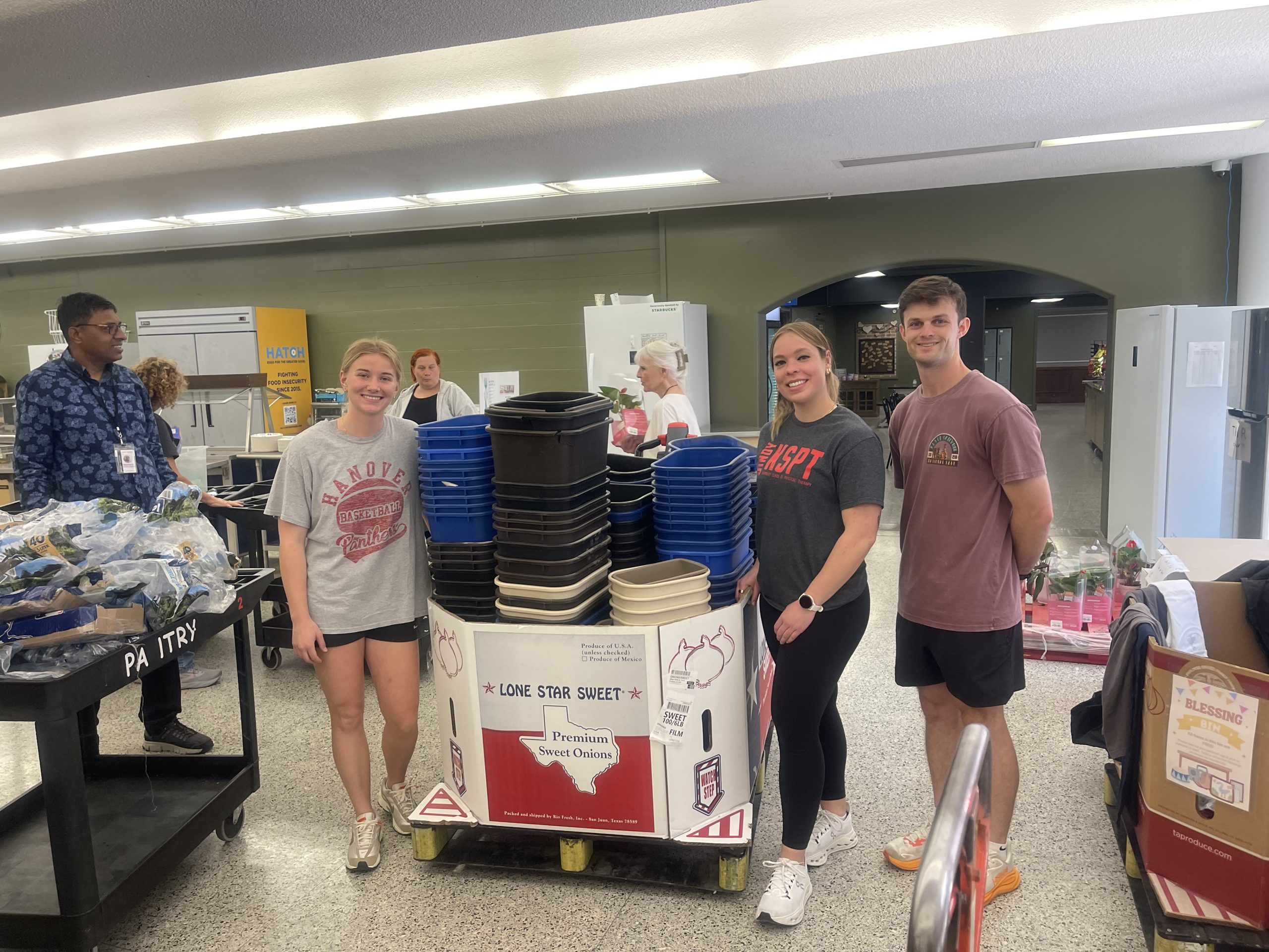 Three students smile with bins at food shelter with adults and stocked items in background