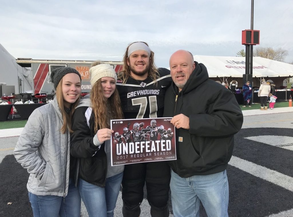 Football player on field in uniform after game holding page that reads "undefeated" is surrounded by family 