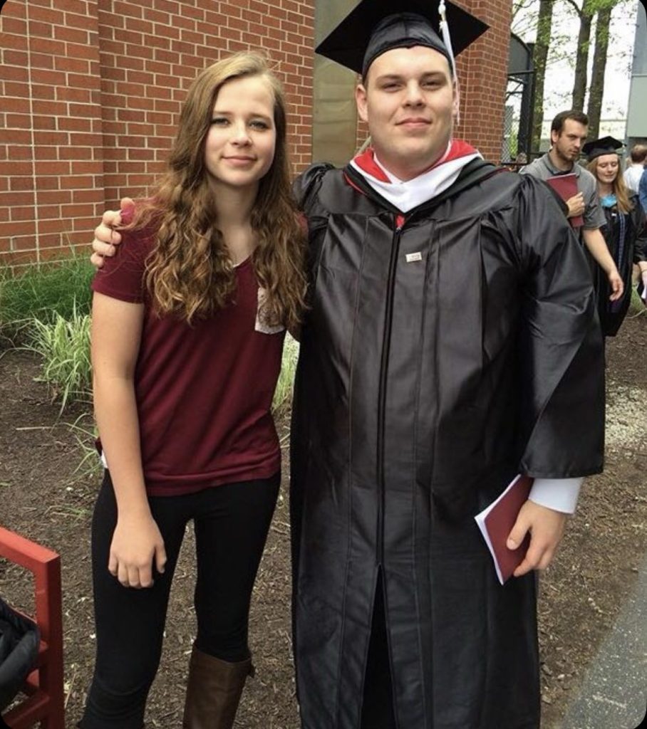 Man in graduation cap and gown has arm around sister with people in background