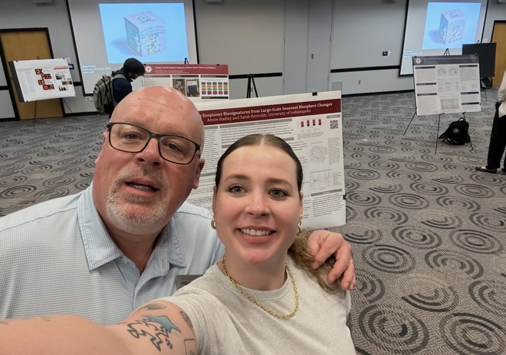 Woman takes selfie with dad's arm wrapped around her. Picture boards and projectors seen in background