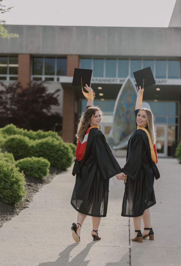 Two girls in graduation gowns hold hands and raise caps to the sky in front of UIndy library
