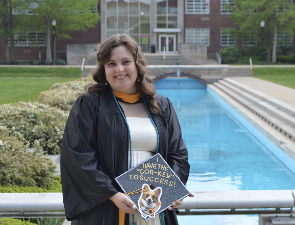 Woman in graduation gown holds graduation cap that says "Have the Cor-Key (Corgie) to success with a picture of corgie. Smith Mall seen in background 