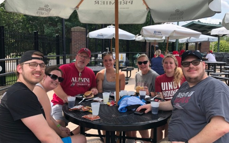 Family sits around public table with burgers and drinks on it and umbrella attached