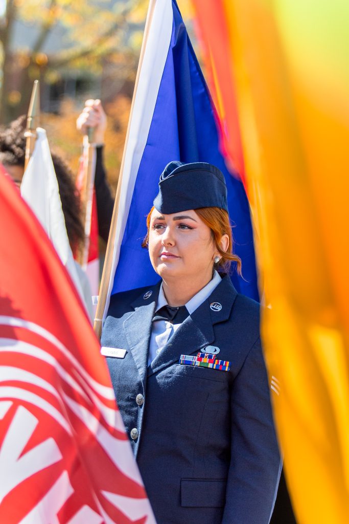 Курсант ROTC стоит по стойке смирно с церемониальным флагом во время мероприятия в кампусе Университета Индианаполиса, обрамленного разноцветными флагами.