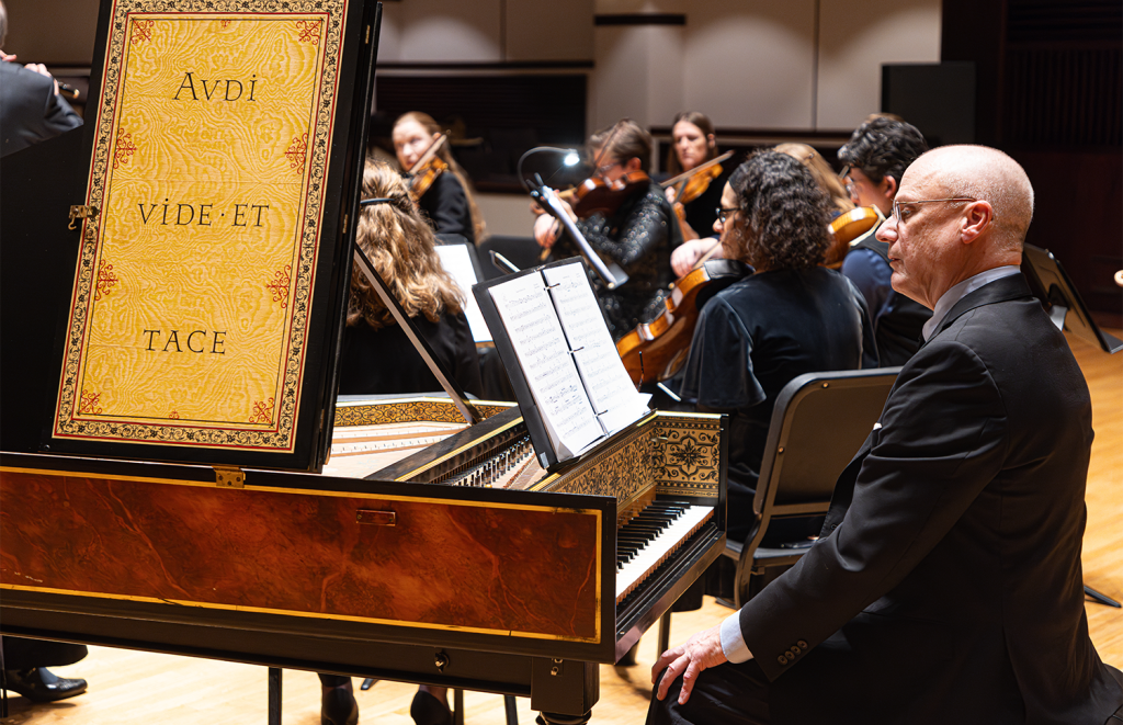 Photo of Tom Gerber, harpsichordist, with the Indianapolis Baroque Orchestra. Text decorating the harpsichord reads "Audi, vide, et tace."
