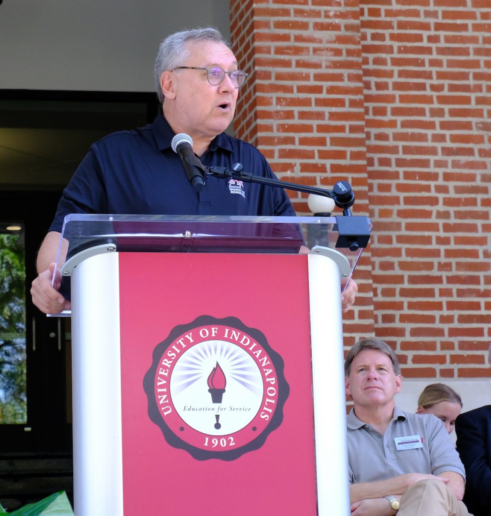 Man stands at podium with UIndy seal and brick wall behind him